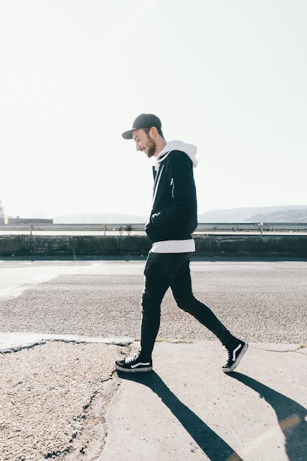 Casual male in a cap and hoodie walks along a sunny urban street.